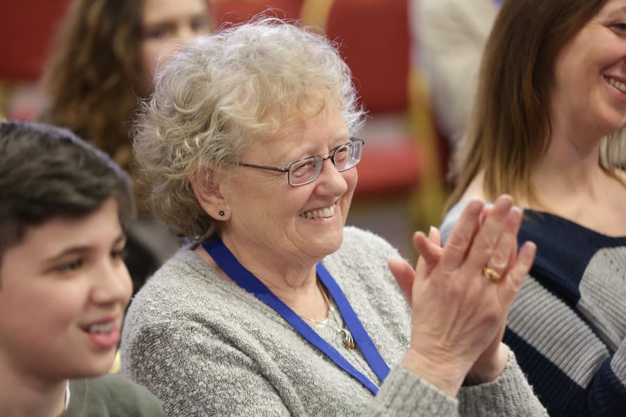 An elderly woman with a big smile clapping during a church conference who's goal is to inspire it's congregation to have a deeper relationship with God.