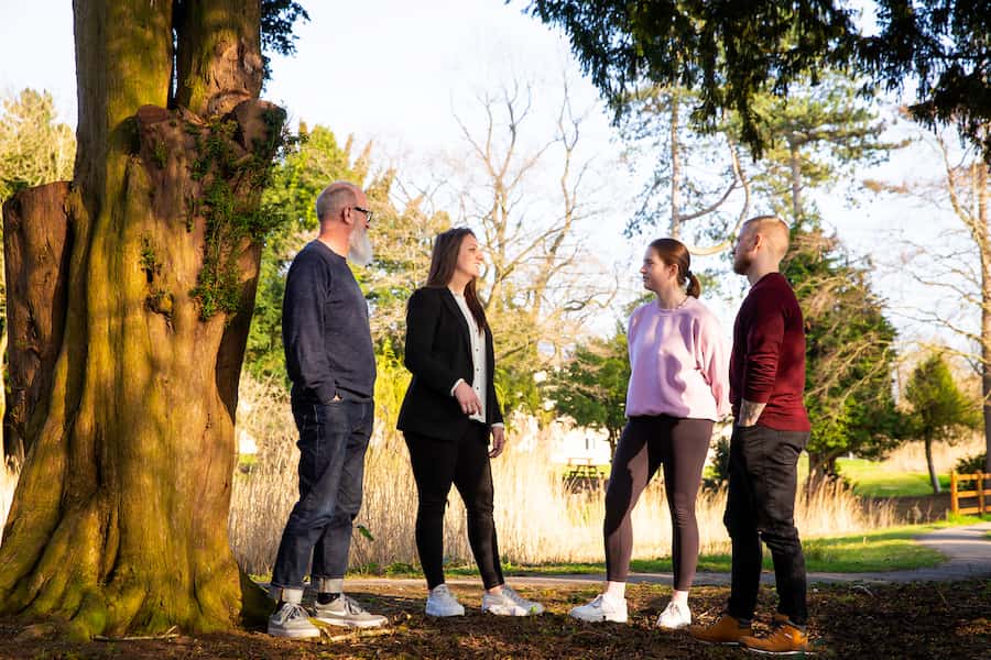 A group of 4 people standing outside besides a big tree discussing ideas of how to help their local community.