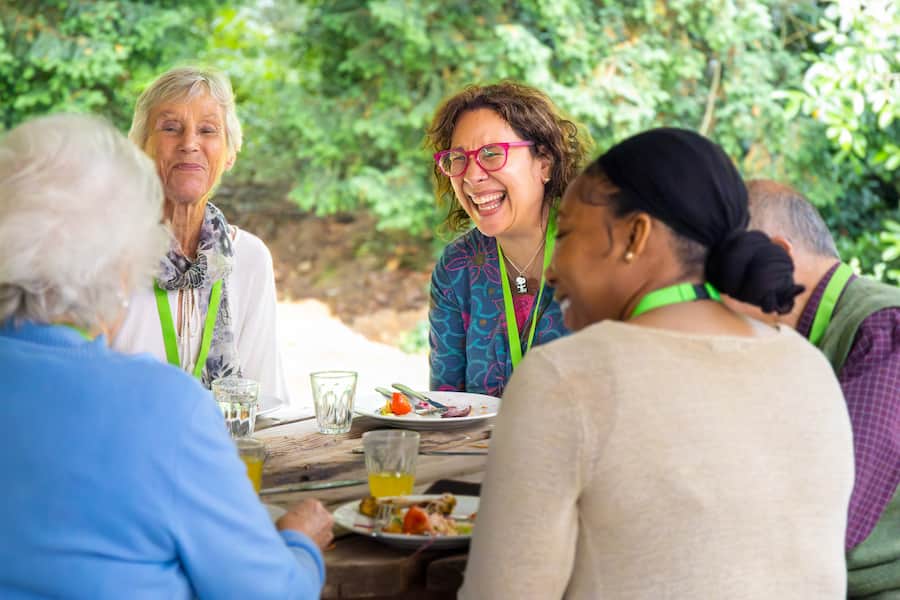 A woman laughing in a group of people around a table outside with food and drink.