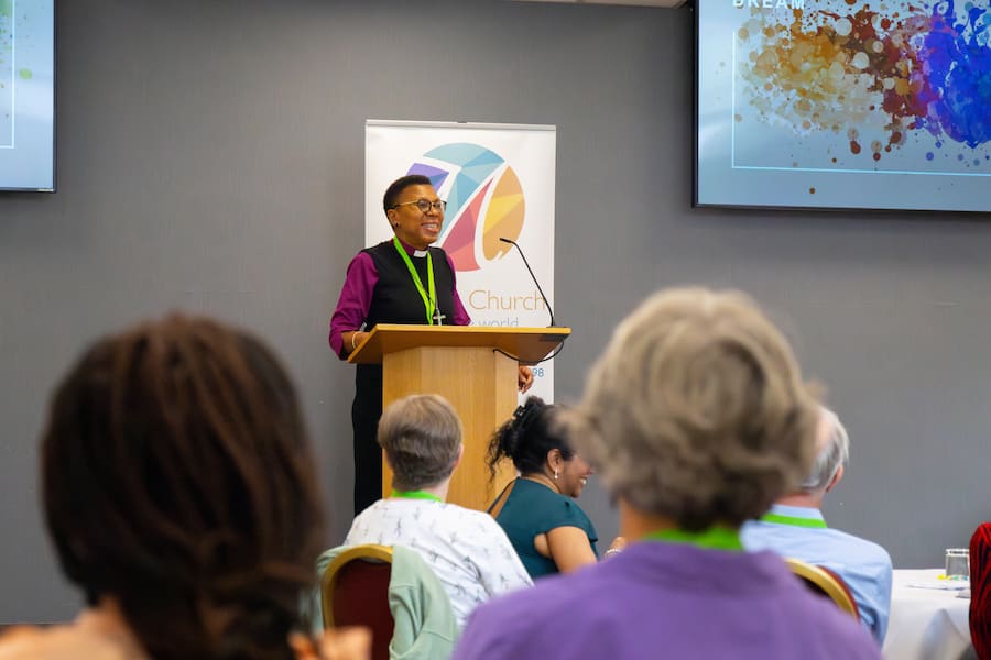 A woman speaking behind a podium at a conference while smiling to a crowd of conference delegates.