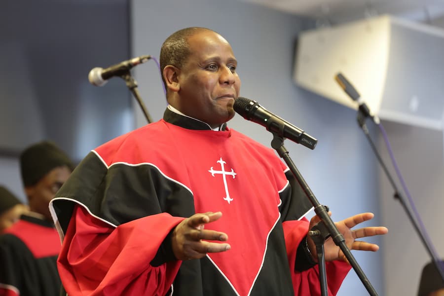 A man speaking into a microphone in red and black dress wear sporting a cross on the front. 