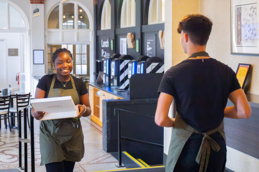 A member of staff passing another member of staff while smiling and carrying tea making facilities.