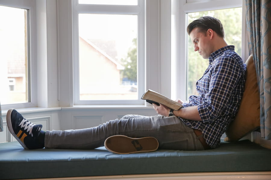 A man sitting with his feet up in a window bay reading the Bible.