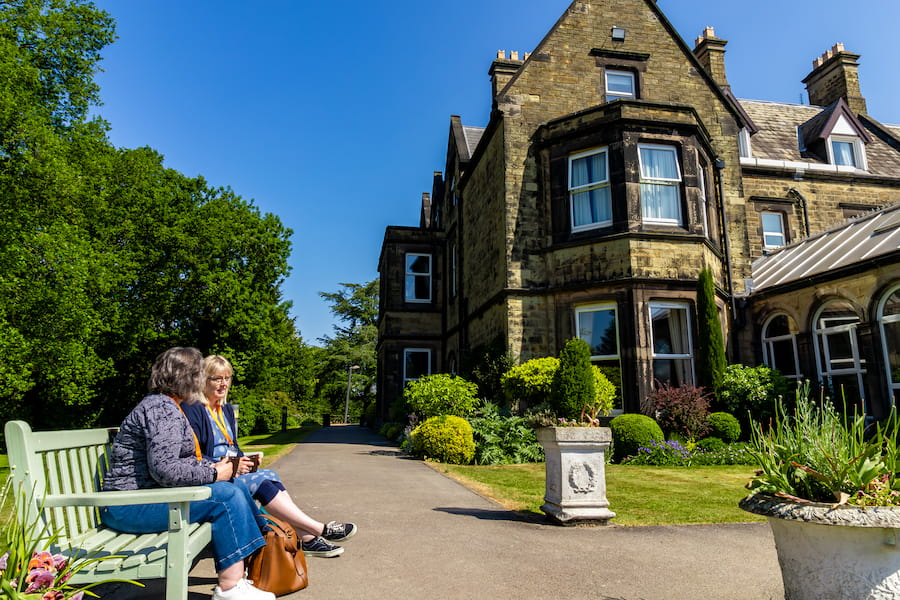 Two women sitting on a bench outside The Hayes in the sunshine enjoying a cup of coffee.